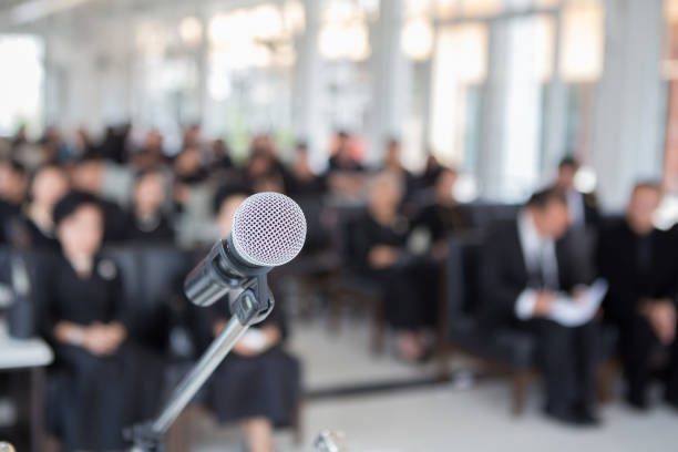 A microphone positioned in front of a conference room, with attendees engaged in discussion in the background
